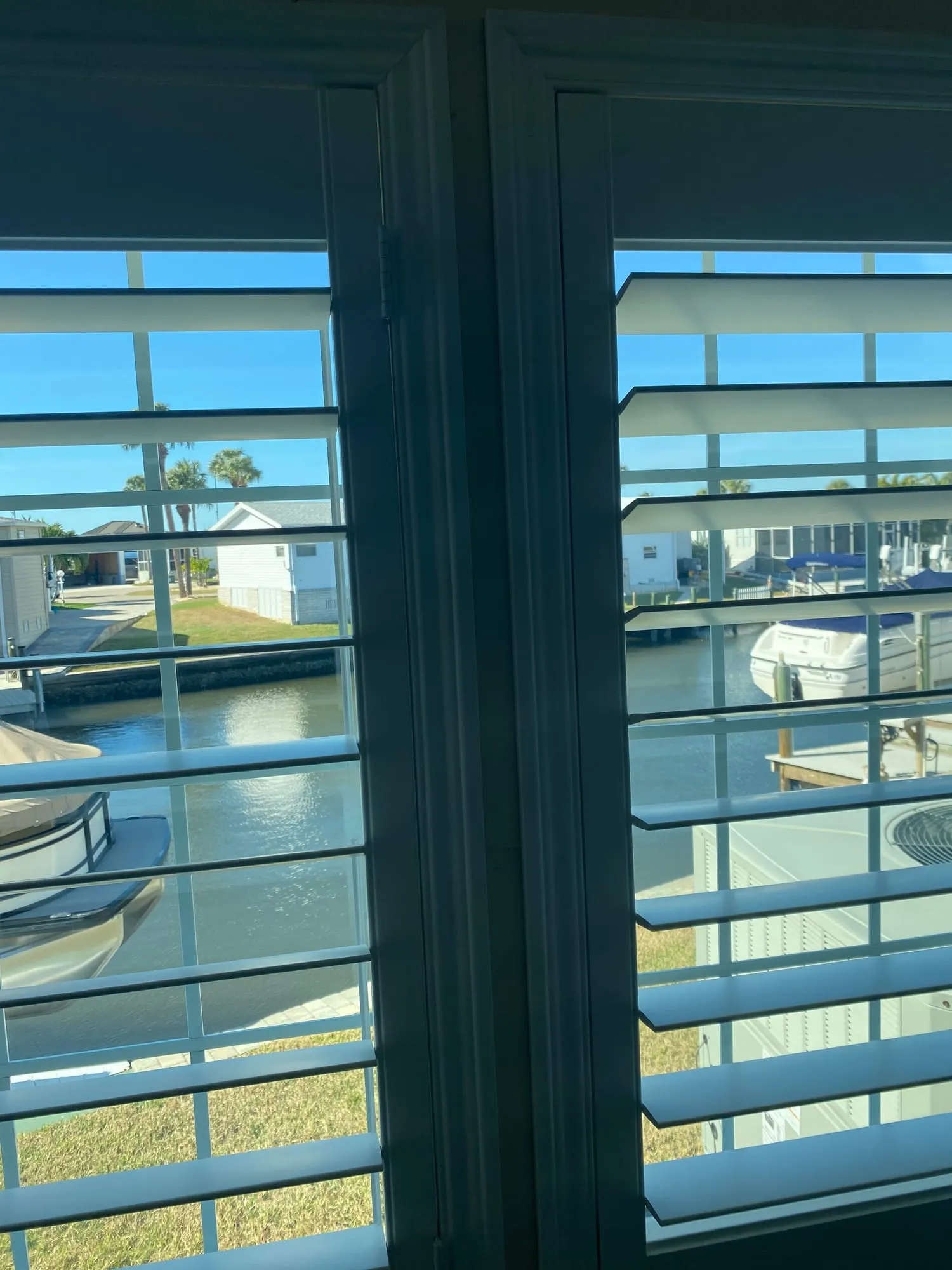 Plantation shutters on a canal-front window, louvers tilted open to boats and palms outside