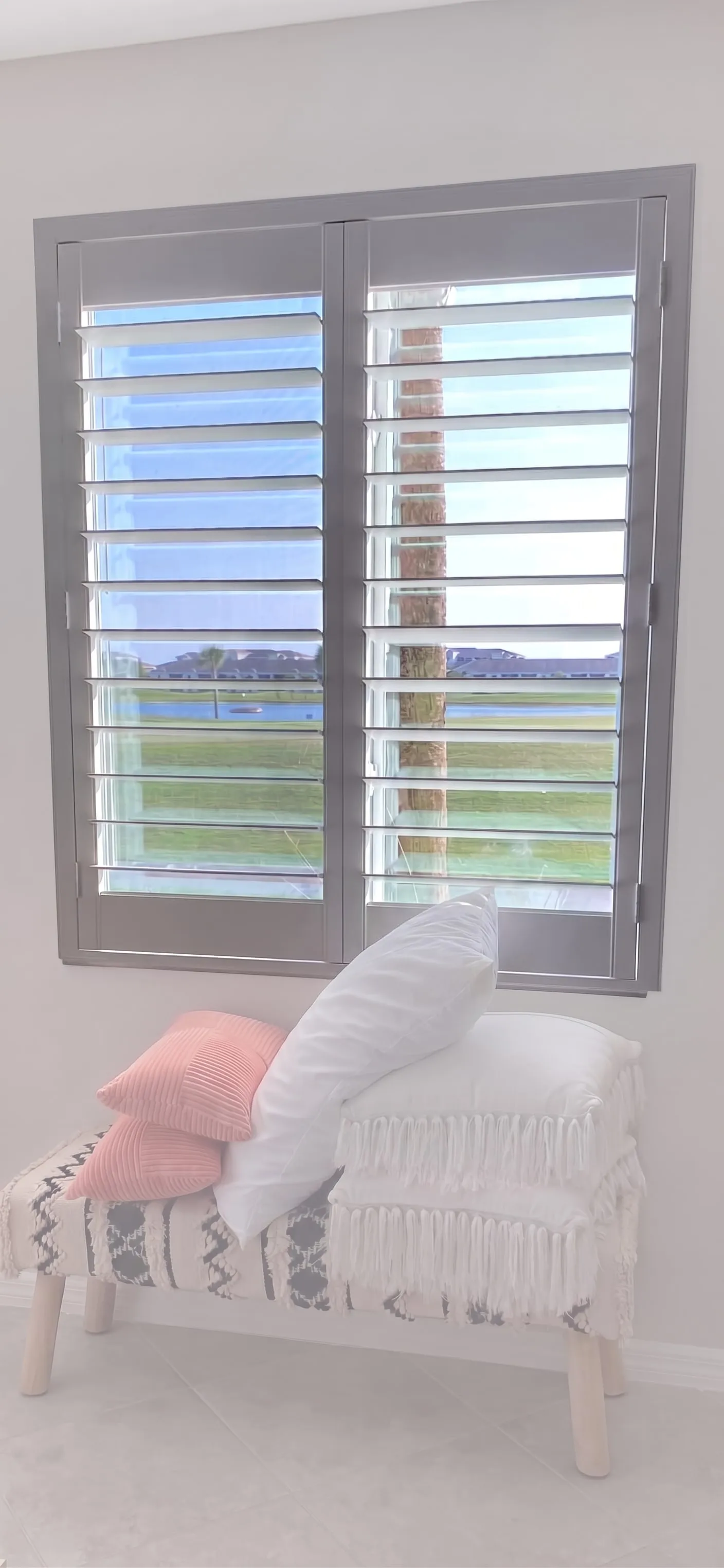 White plantation shutters with louvers open to a lake view, above a bench with throw pillows in a Southwest Florida home